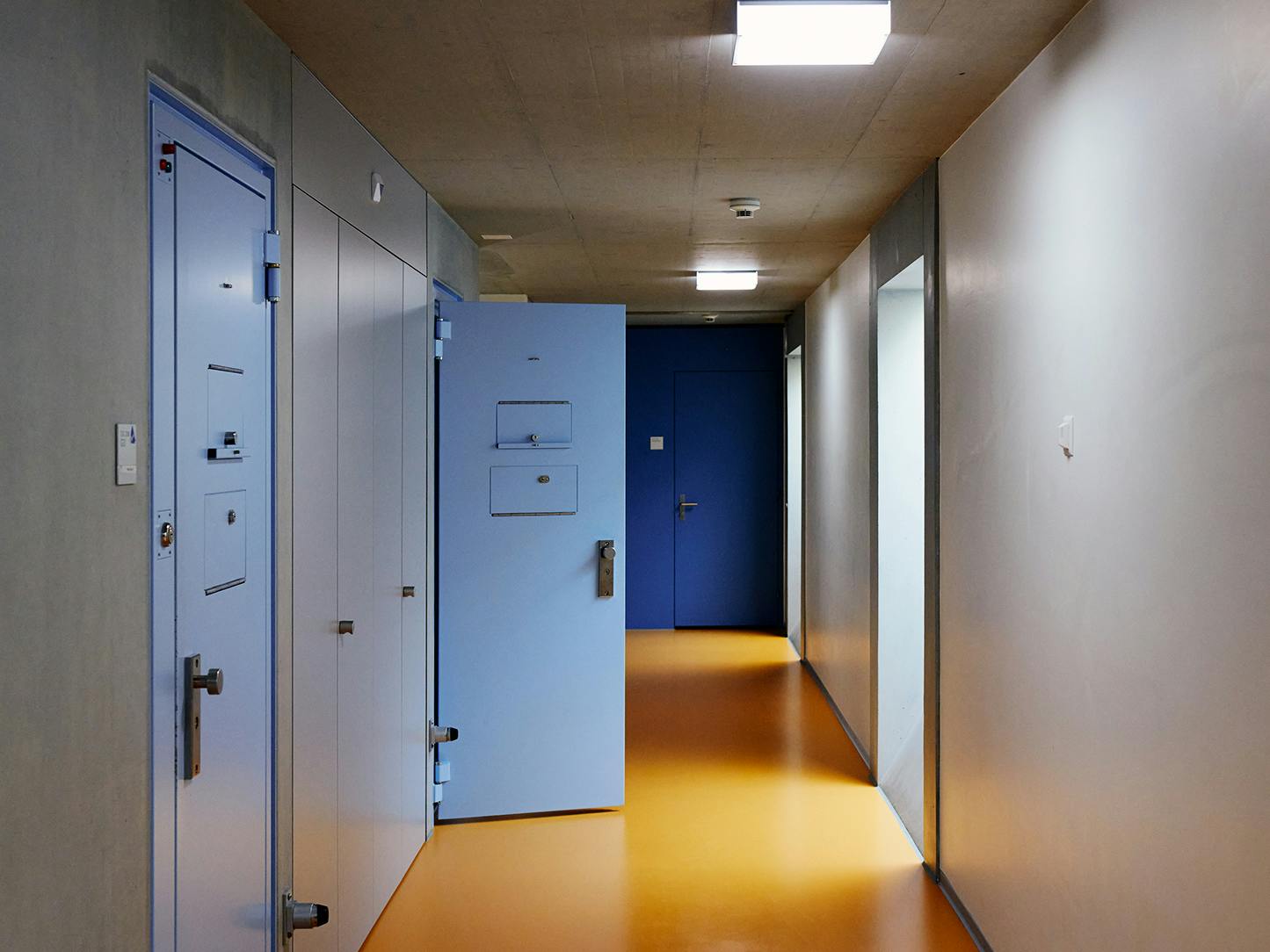 Gefängniskorridor mit gelben Boden und hellblauen Türen.