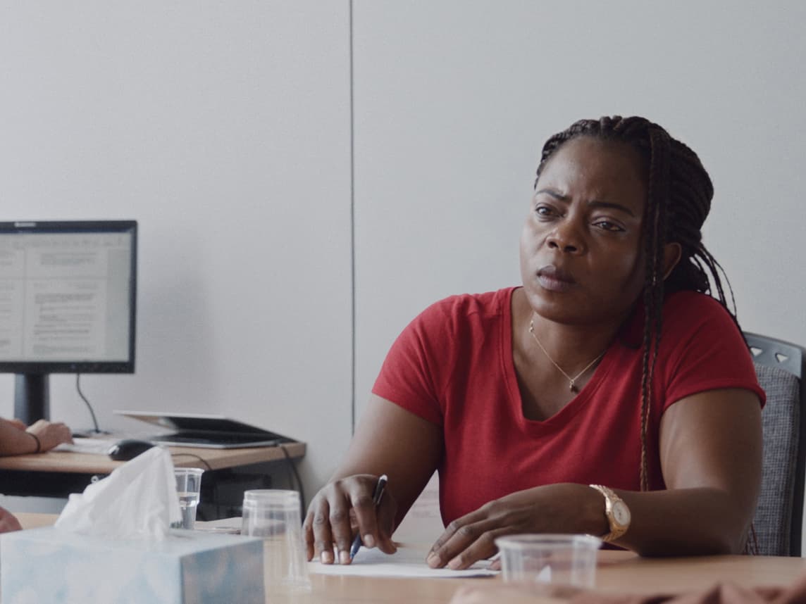 Filmstill Die Anhörung. Frau mit rotem Tshirt sitzt an einem Tisch